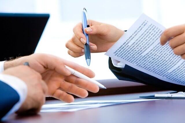 Imagen que enfoca las manos de dos personas, una tine un bolígrafo azul en la mano derecha y un documento en la izquierda; el otro tiene un bolígrafo blanco en la mano izquierda señalando unos papeles sobre el escritorio.